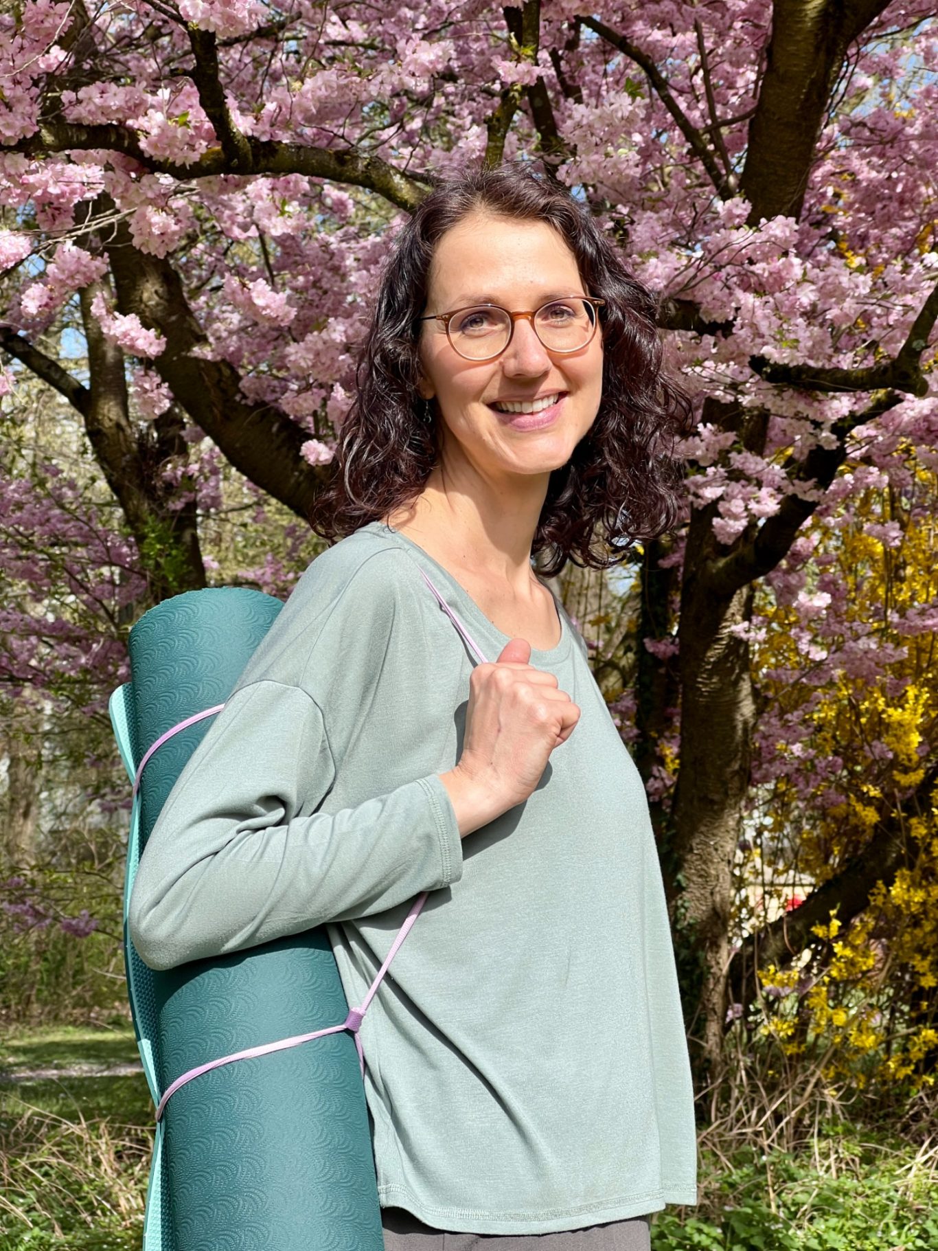 Frau mit Yogamatte vor blühenden Kirschbäumen, lächelnd in der Natur.