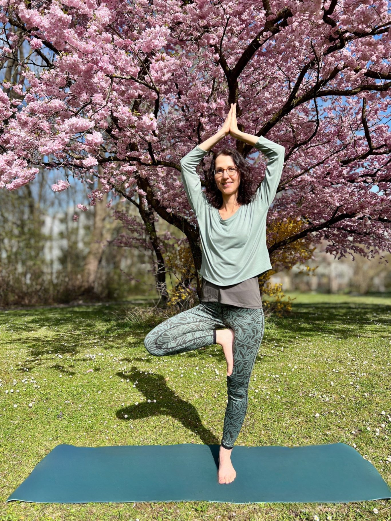 Eine Frau in Yoga-Pose auf einem grünen Rasen vor blühenden Kirschbäumen.